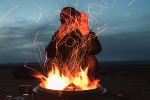 Person warming their hands by the embers of flames of a campfire at dusk.