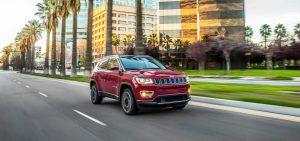 A red 2021 Jeep Compass driving down a city road on a sunny day, with palm trees and apartment complexes in the background.