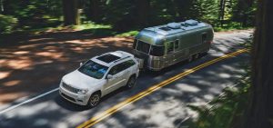 White Jeep Grand Cherokee towing a trailer behind it on a sunny day beneath a canopy of trees.