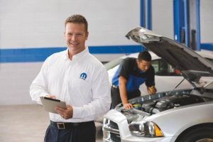 Service man holding a clipboard while smiling. Another man working under the hood of a car.